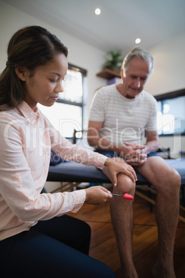 Female therapist examining knee of senior male patient with reflex hammer