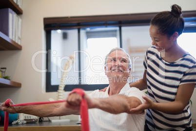 Smiling senior male patient pulling red resistance band while looking at female doctor