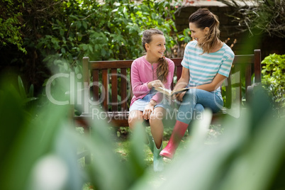 Happy mother and daughter looking at each other while sitting with novel on bench