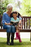 Smiling grandmother and granddaughter reading novel while sitting on wooden bench