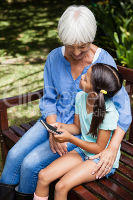 Granddaughter sitting with mobile phone looking at grandmother on wooden bench