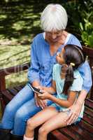 Granddaughter sitting with mobile phone looking at grandmother on wooden bench