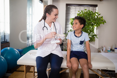 Smiling young female therapist looking at boy with artificial bone while sitting on bed
