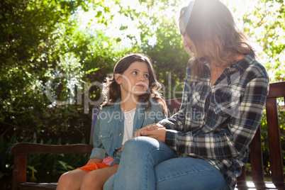 Girl sitting with mother on wooden bench against trees during sunny day