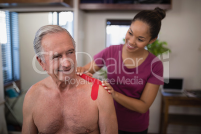 Female therapist applying elastic therapeutic tape on shoulder of smiling shirtless senior male pati