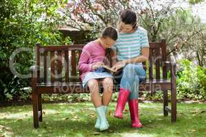 Mother and daughter reading novel while sitting on wooden bench