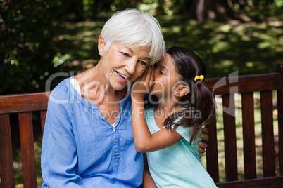 Girl whispering in ears of grandmother while sitting on wooden bench