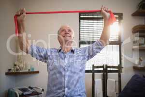 Smiling senior male patient pulling red resistance band while looking up