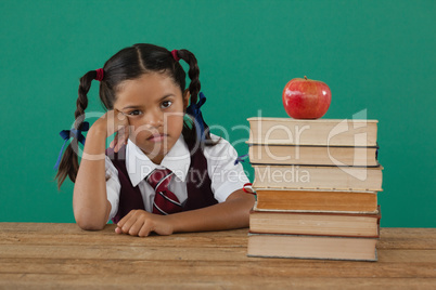 Schoolgirl sitting beside books stack with apple on top against chalkboard