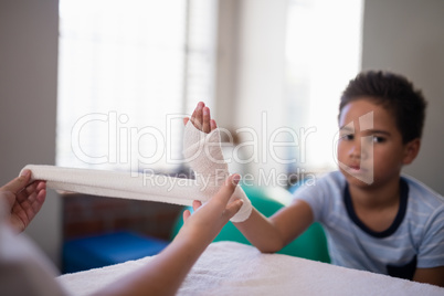 Portrait of boy sitting at table while female therapist wrapping bandage on hand