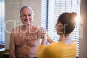 Smiling shirtless senior male patient looking at female therapist giving arm massage