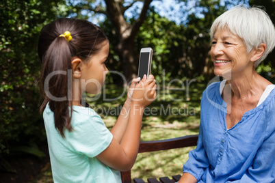 Side view of girl taking photograph of grandmother sitting on bench