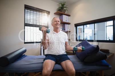 Smiling senior male patient lifting dumbbells while sitting on bed