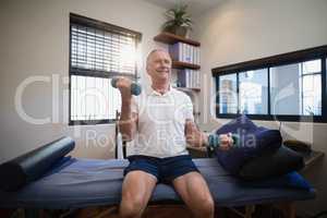 Smiling senior male patient lifting dumbbells while sitting on bed