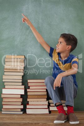 Schoolboy sitting on stack of book in front of chalkboard in classroom