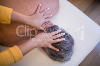 Rear view of shirtless senior male patient lying on bed receiving neck massage from female therapist