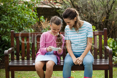 Smiling woman sitting by girl using mobile phone on wooden bench