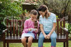 Smiling woman sitting by girl using mobile phone on wooden bench