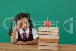 Unhappy schoolgirl looking at books stack and apple against chalkboard