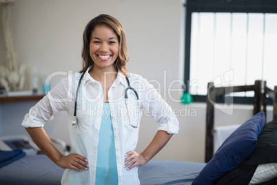 Portrait of smiling female therapist standing with hands on hip by bed