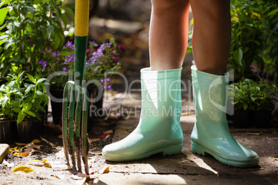 Low section of girl wearing green rubber boot standing with gardening fork on footpath