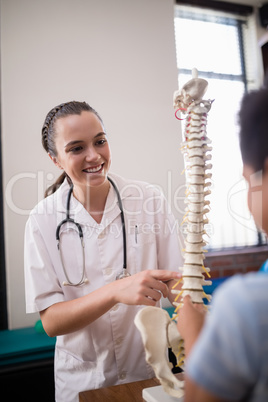 Smiling female therapist explaining boy while pointing at artificial spine against wall