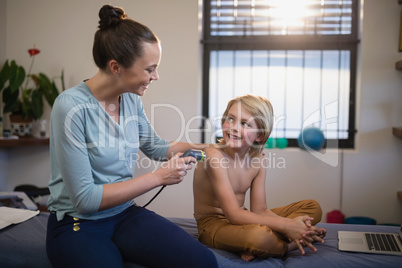 Smiling young female therapist scanning shoulder while looking at shirtless boy sitting on bed