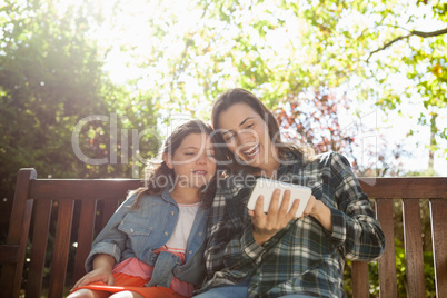 Cheerful woman showing mobile phone to daughter while sitting on wooden bench