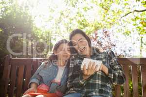 Cheerful woman showing mobile phone to daughter while sitting on wooden bench