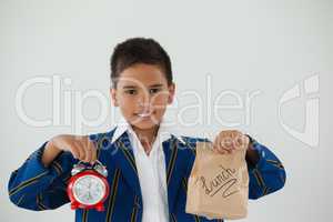 Schoolboy holding alarm clock and disposable lunch bag against white background