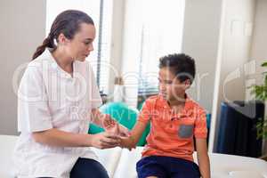 Young female therapist examining hand of boy while sitting on bed