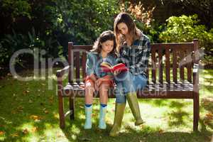 Woman reading book to daughter while sitting on wooden bench