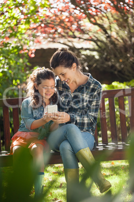 Smiling girl and mother using mobile phone while sitting on wooden bench