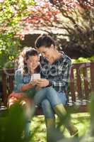 Smiling girl and mother using mobile phone while sitting on wooden bench