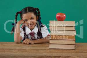 Schoolgirl sitting beside books stack with apple on top against chalkboard