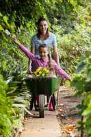 Woman pushing girl sitting in wheelbarrow on footpath amidst plants