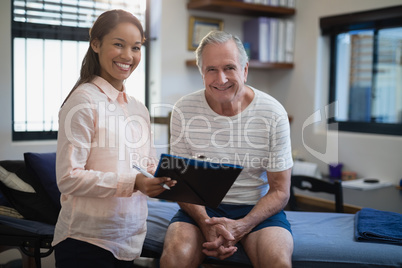 Portrait of smiling female therapist standing with clipboard by senior male patient sitting on bed