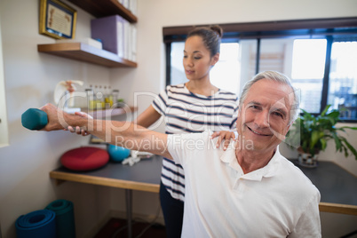 Female doctor holding hand of smiling senior male patient lifting dumbbell