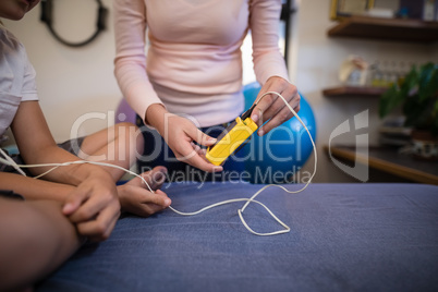 Midsection of female therapist showing electrical muscle stimulation machine to boy sitting on bed
