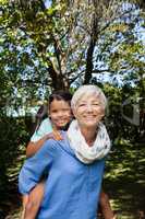 Portrait of smiling grandmother giving piggyback to granddaughter against trees