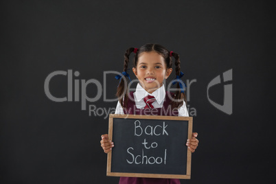 Portrait of schoolgirl holding slate with text against blackboard