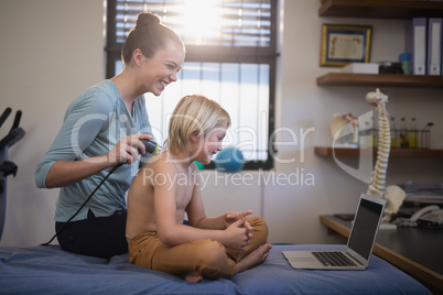 Cheerful female therapist while scanning shoulder of shirtless boy looking at laptop