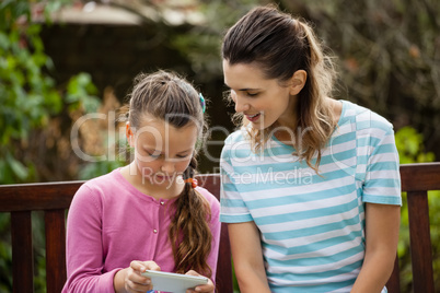 Woman sitting by girl using smartphone on wooden bench