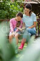 Girl sitting with mother reading novel on wooden bench