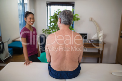 Rear view of shirtless senior male patient sitting on bed looking at smiling female therapist