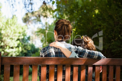 Rear view of mother and daughter sitting with arms around on wooden bench
