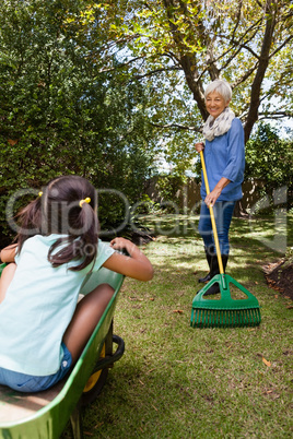 Grandmother holding rake while looking granddaughter sitting in wheelbarrow