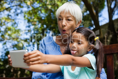Grandmother and granddaughter making face while taking selfie sitting on bench