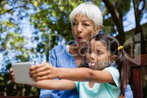 Grandmother and granddaughter making face while taking selfie sitting on bench