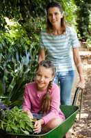 Happy mother pushing girl with flowers sitting in wheelbarrow